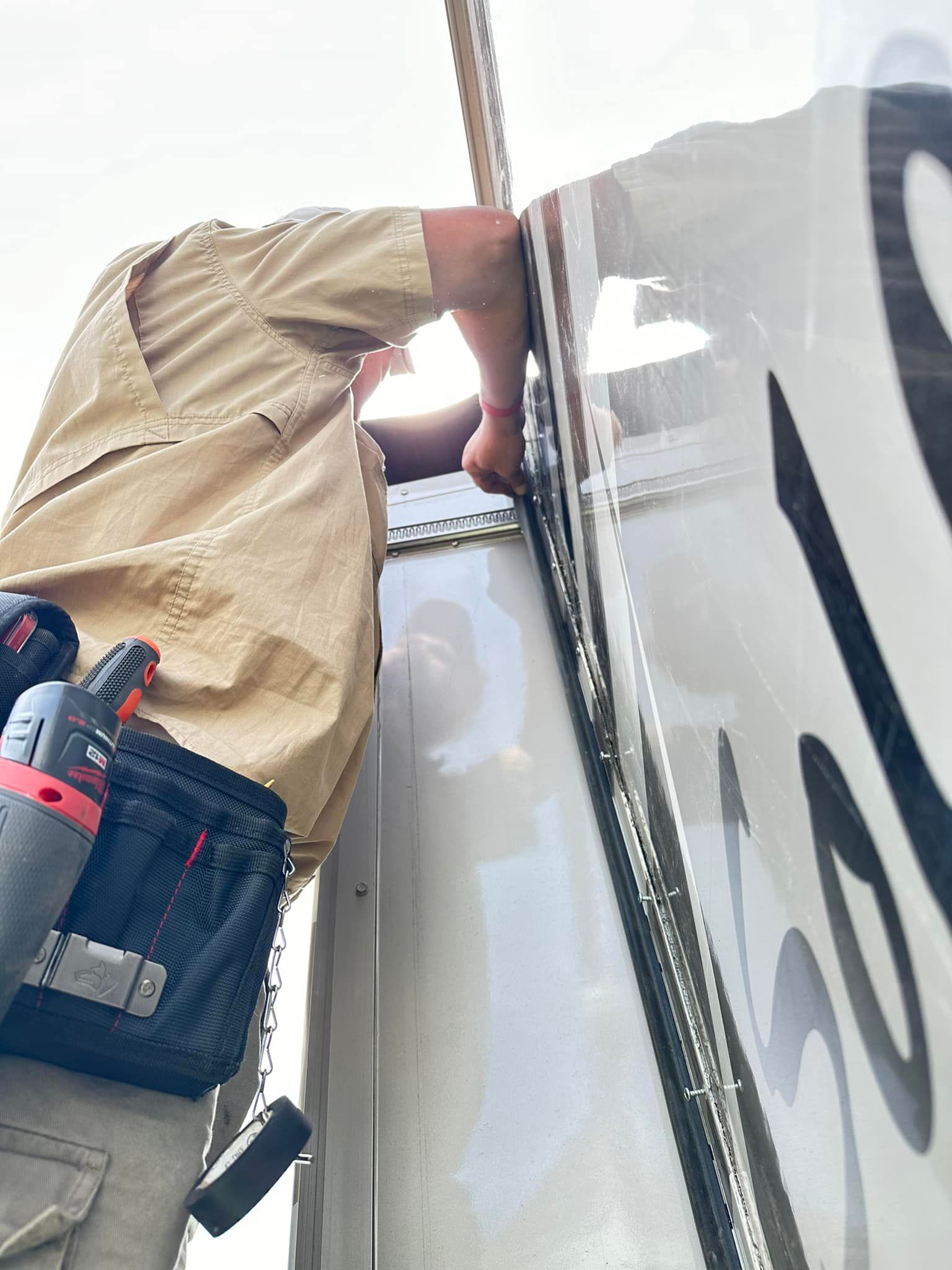 Person in tan shirt, working on a white vehicle, holding something near its top. A tool belt is visible.