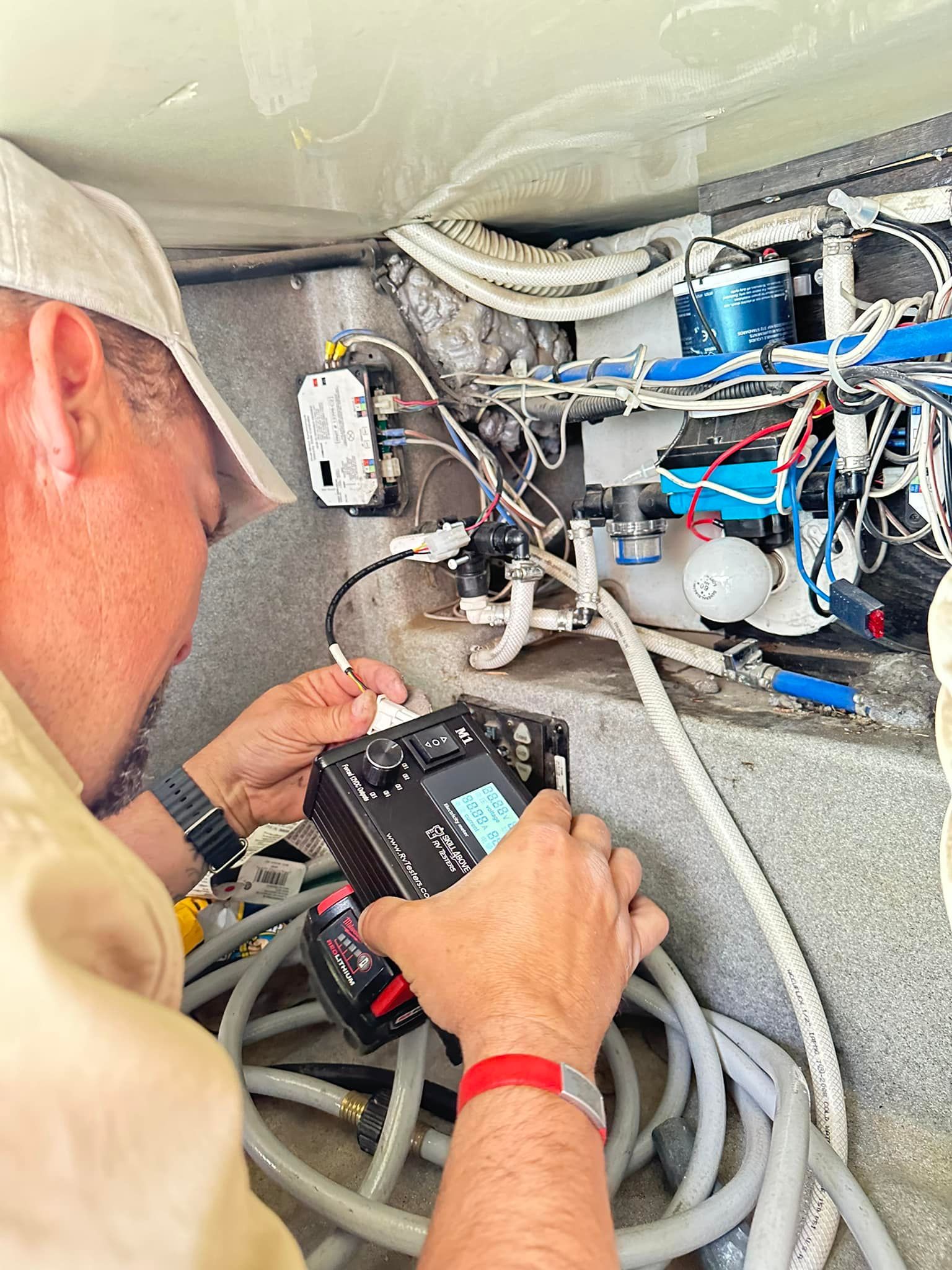 Person in a cap using a black testing device inside a complex system of wires, pipes, and other components.