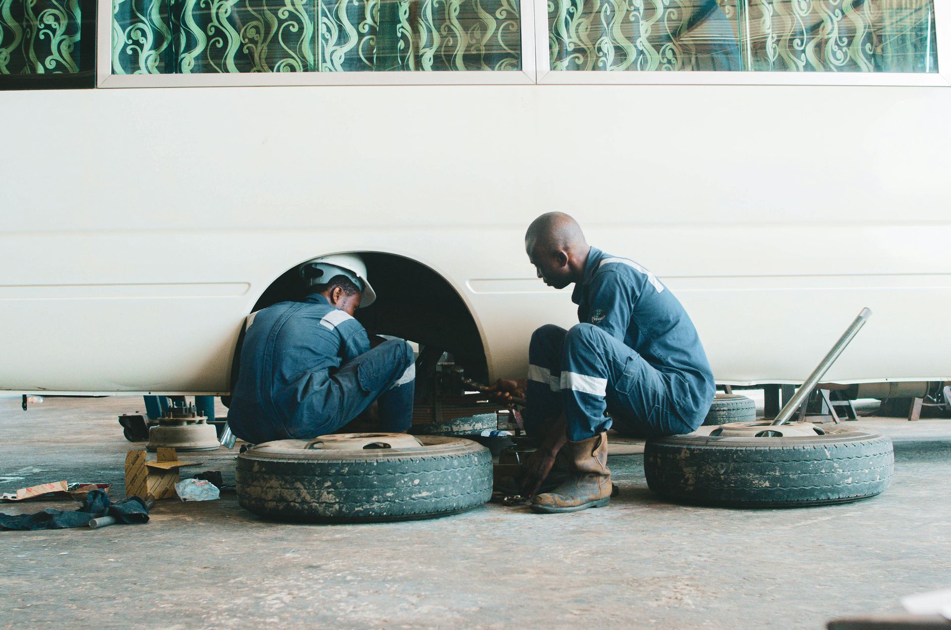 Two mechanics in blue work uniforms repairing the wheel assembly of a large white bus inside a garage.