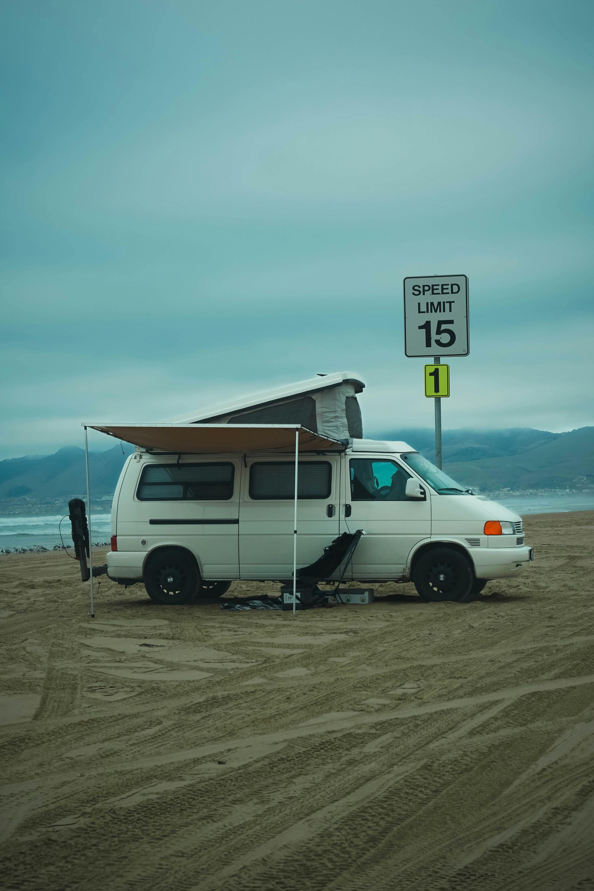 White camper van parked on a sandy beach under a cloudy sky near a speed limit sign.