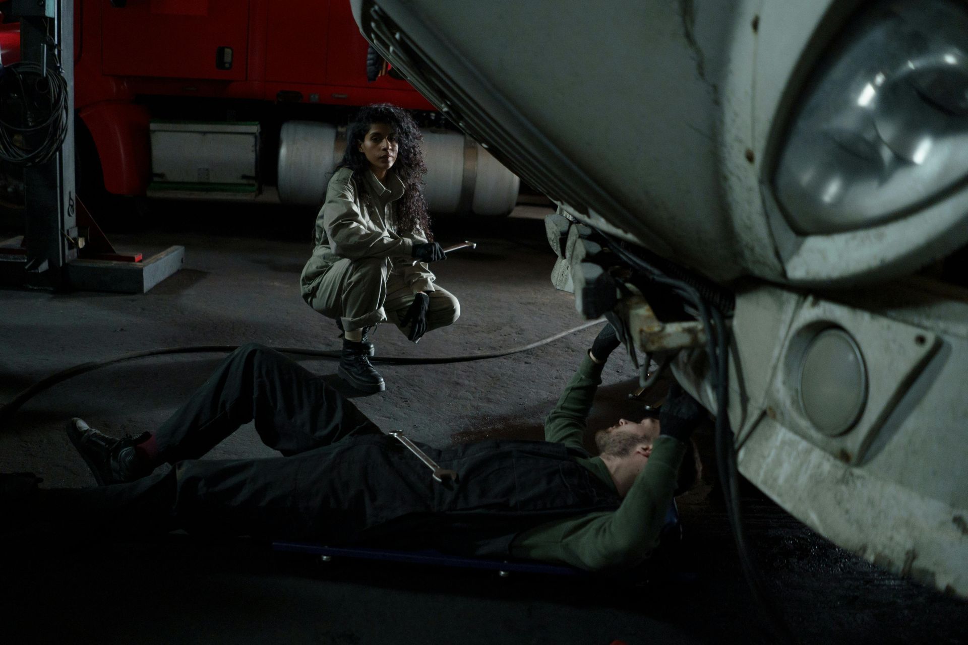 Two people repairing the underside of a large white vehicle in a dimly lit garage or parking area.