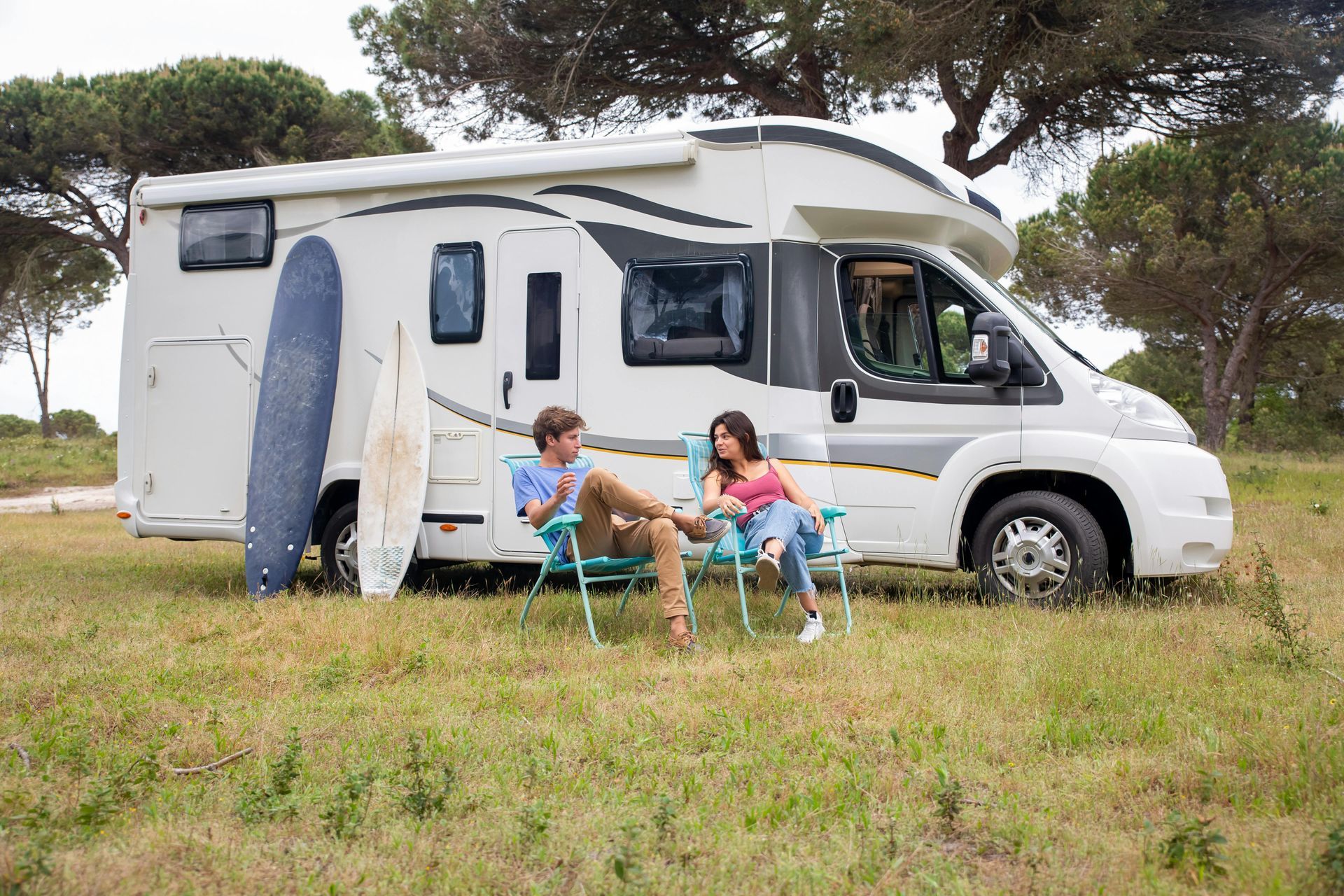 Couple seated in chairs next to a white RV, surfboard leaning against it, outdoors in a grassy area.