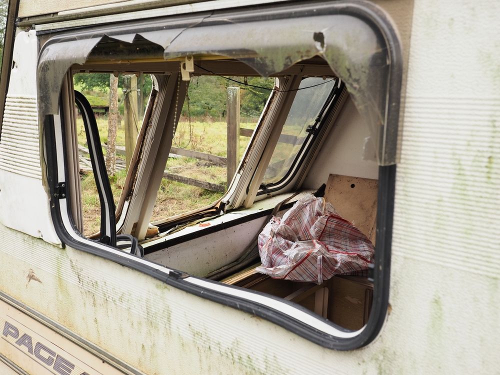 Damaged caravan window; broken plastic, interior visible with debris. Outdoors, overcast day.