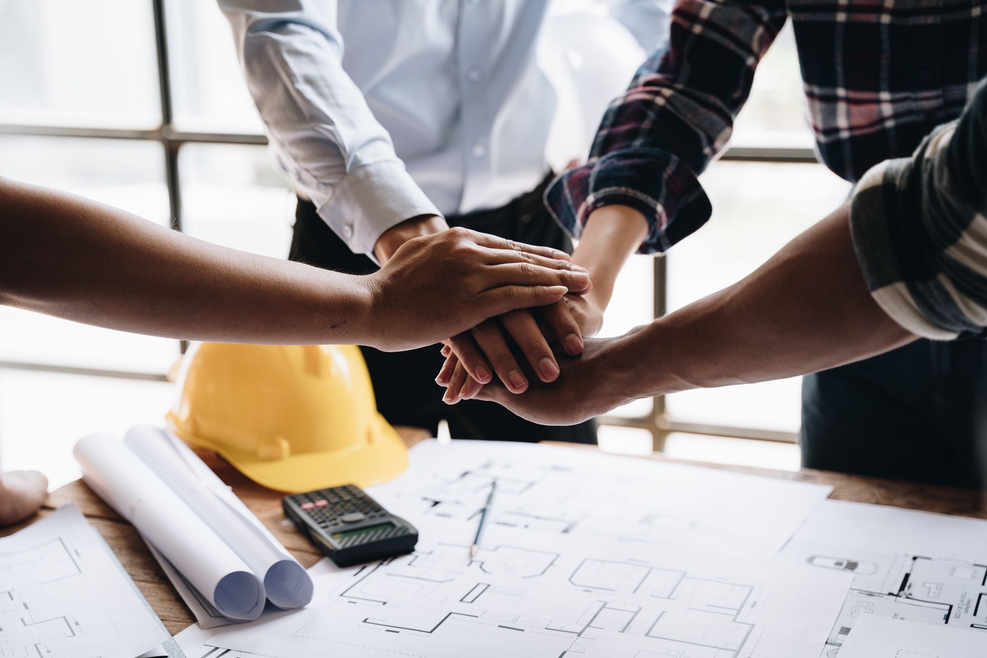 Hands of a team stacked together over blueprints and a yellow hard hat on a table, signifying unity and teamwork.