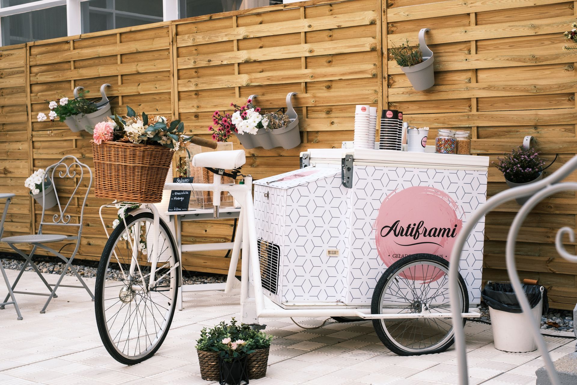 White ice cream cart with bicycle, flowers, and wooden fence background.