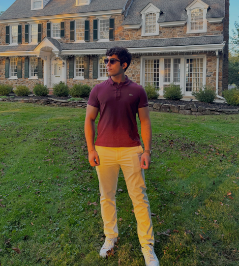 Man in burgundy polo and white pants standing on a lawn in front of a large house.