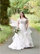 Bride in white tiered wedding dress holding bouquet, standing on path, trees in background.