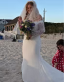 Bride in white dress waves, holding bouquet, on beach with child.
