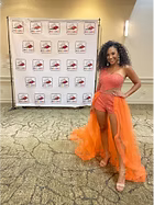 Woman in orange formal wear poses in front of a banner.