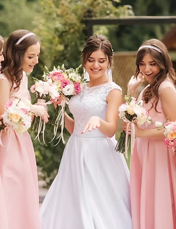 Bride and bridesmaids in pink dresses, looking at a ring, holding bouquets, in a garden.