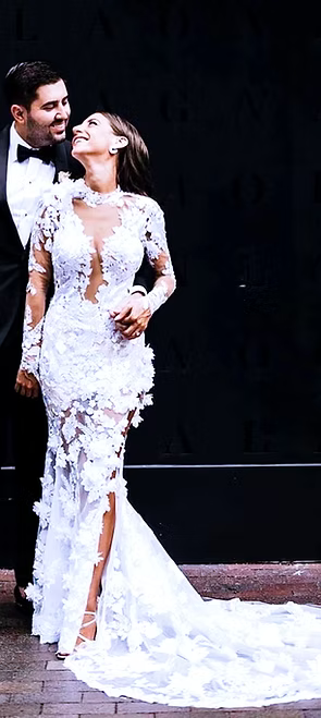 Couple gazing at each other. Bride in white, floral lace dress with long train. Groom in black suit. Black background.
