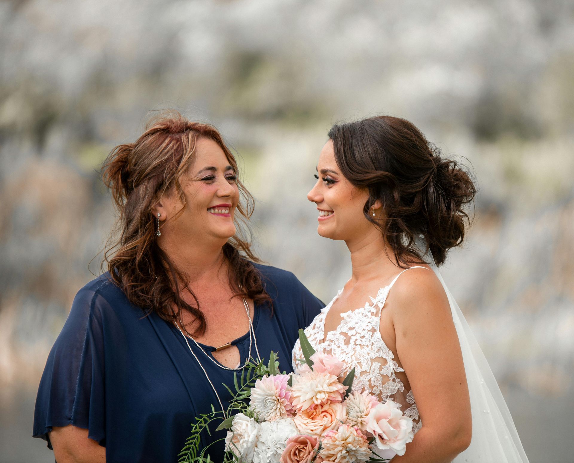 Bride in white gown embraces woman in pink dress, smiling near a floral backdrop.