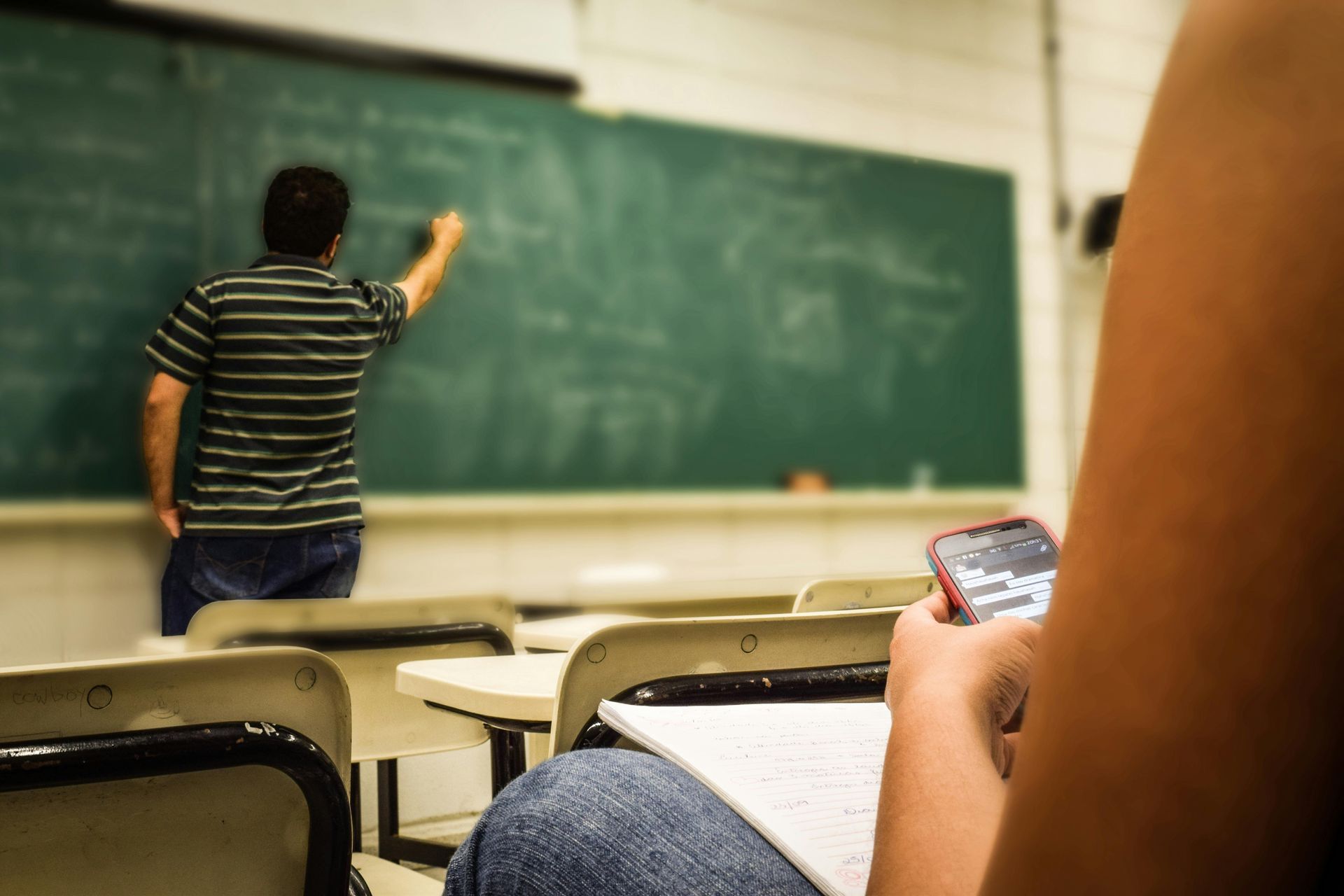Student in a classroom texting while the teacher writes on the chalkboard.
