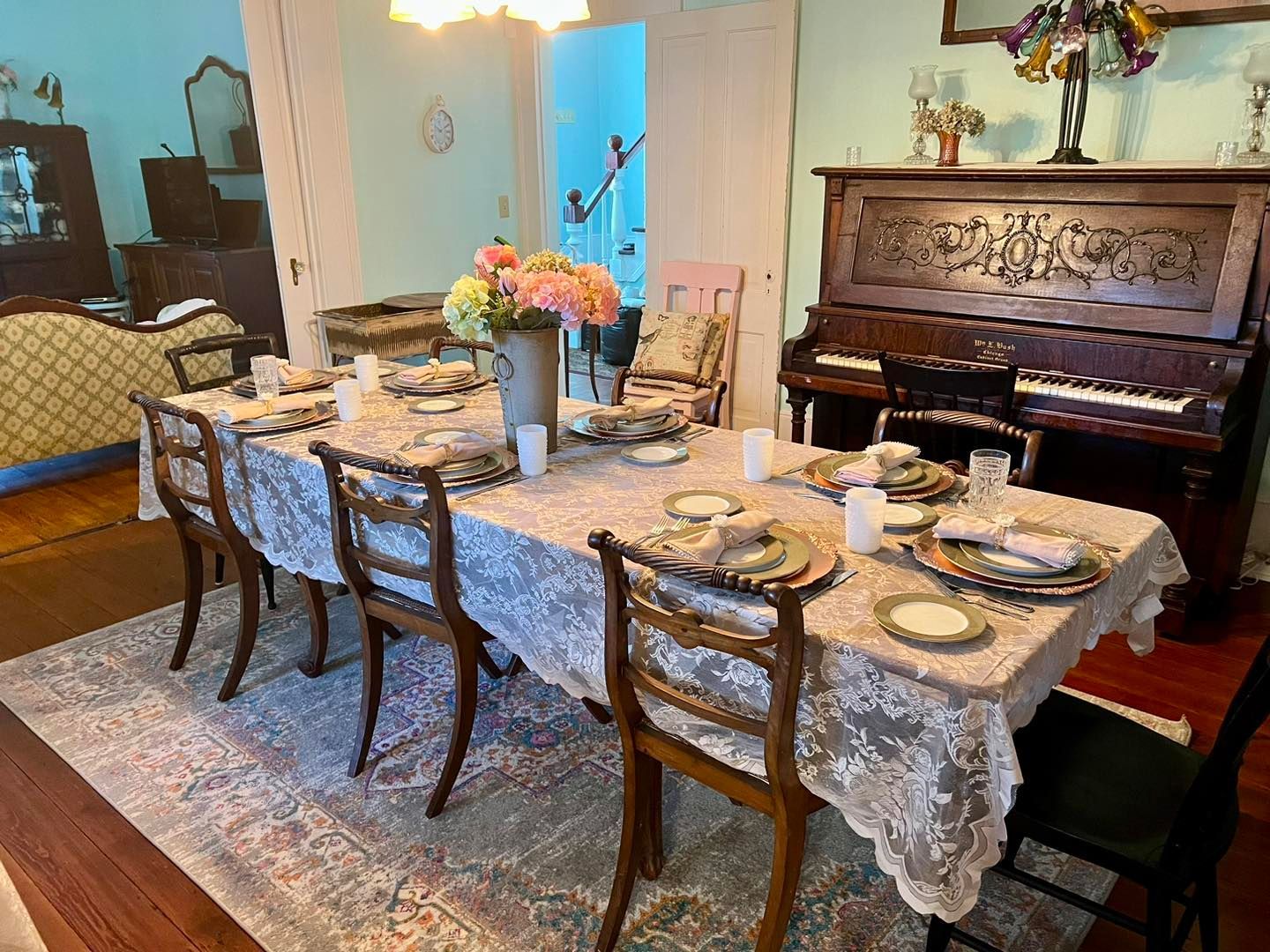 A long table with a lace tablecloth and chairs in a dining room with a piano.