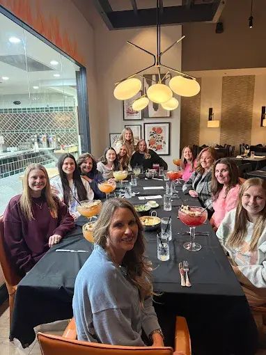 Group of people at a restaurant table. They are sitting around a table with drinks, smiling, in a brightly lit setting.