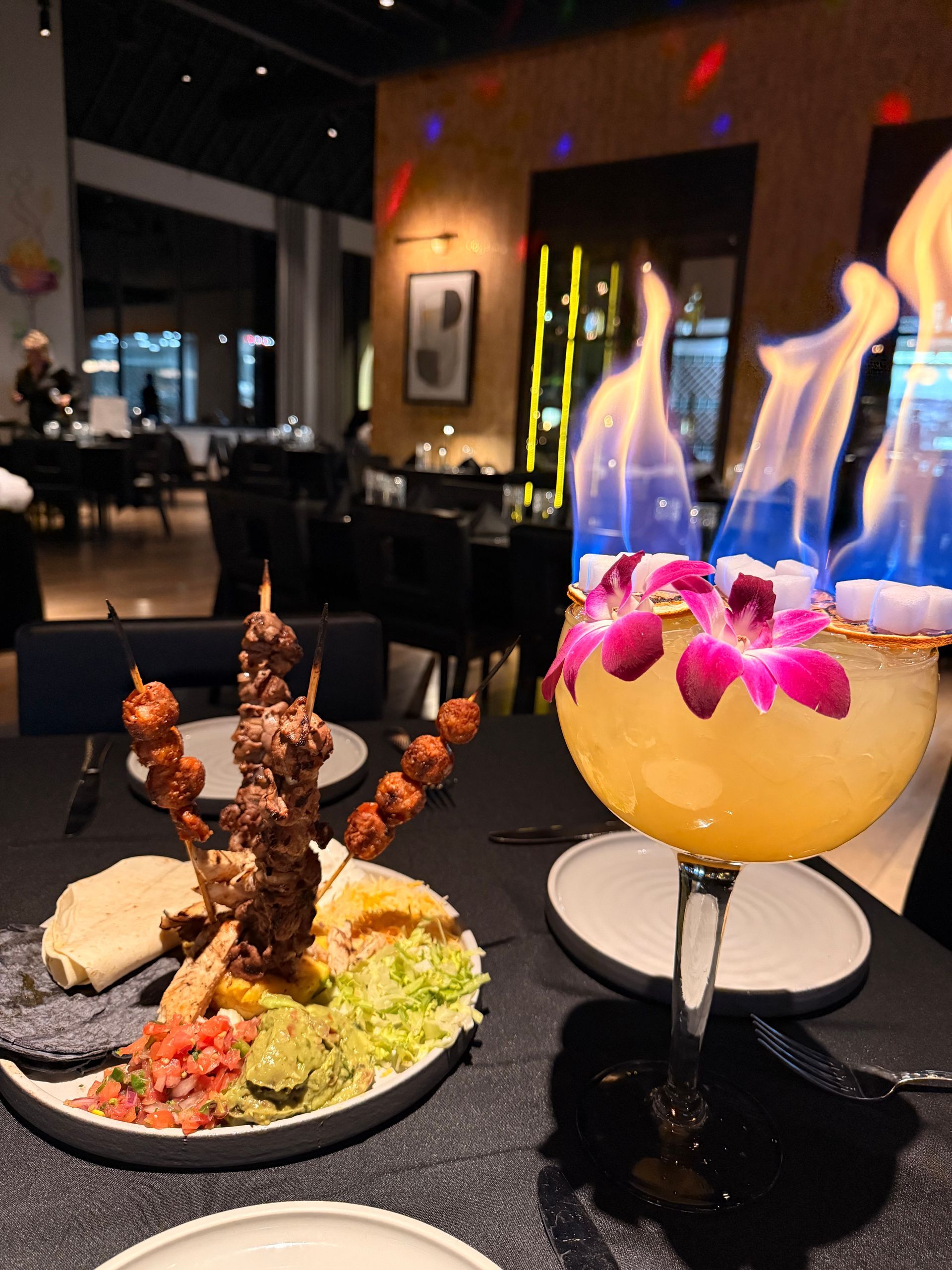 Flame-topped cocktail and food platter on a table at a restaurant. Skewered meat and dipping sauces are visible.