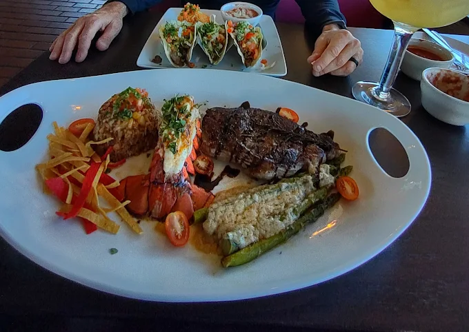 Plate with steak, lobster tail, stuffed corn, tacos, and margarita. Person's hands present.