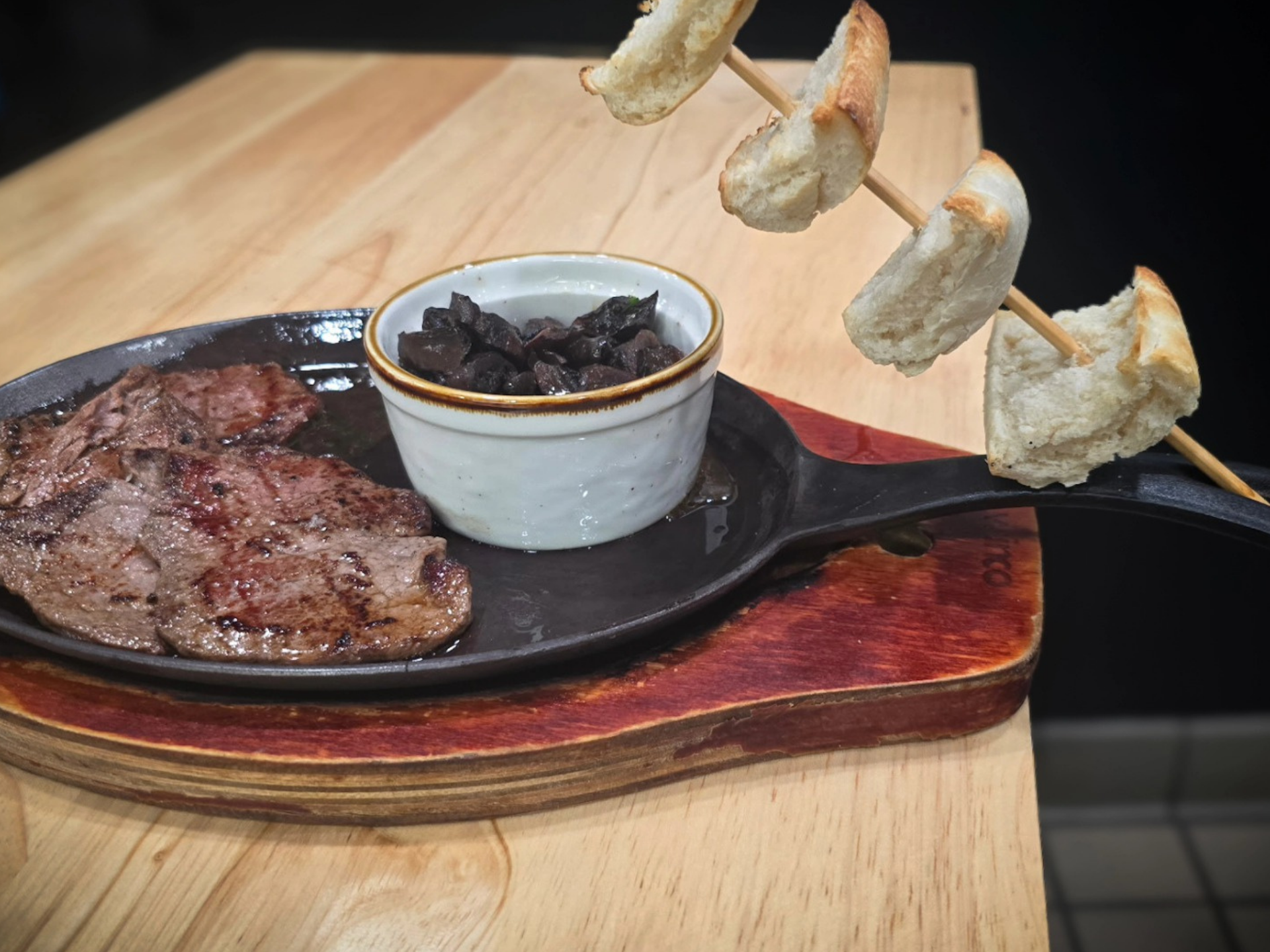 Steak on a cast iron plate with a side of black olives and bread skewers on a wooden surface.