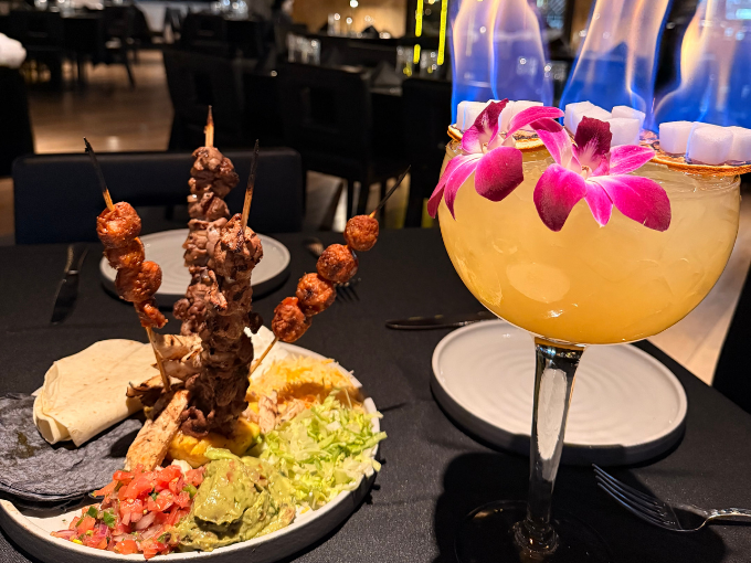 A fancy meal with skewers and a large cocktail garnished with a flower. The drink is set aflame.