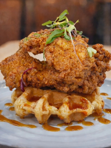 Fried chicken stacked on a waffle, drizzled with sauce, garnished with sprouts, on a gray plate.