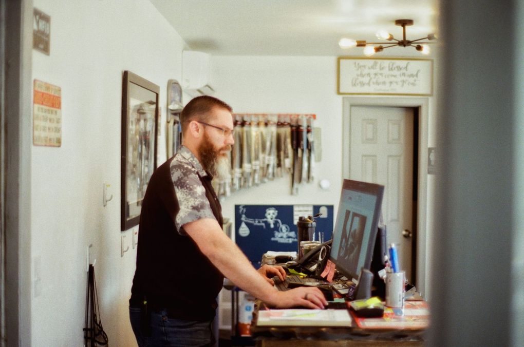 Man standing at a cluttered desk in a bright hallway, using a computer with tools and framed art nearby. | Buck & Doe Auto