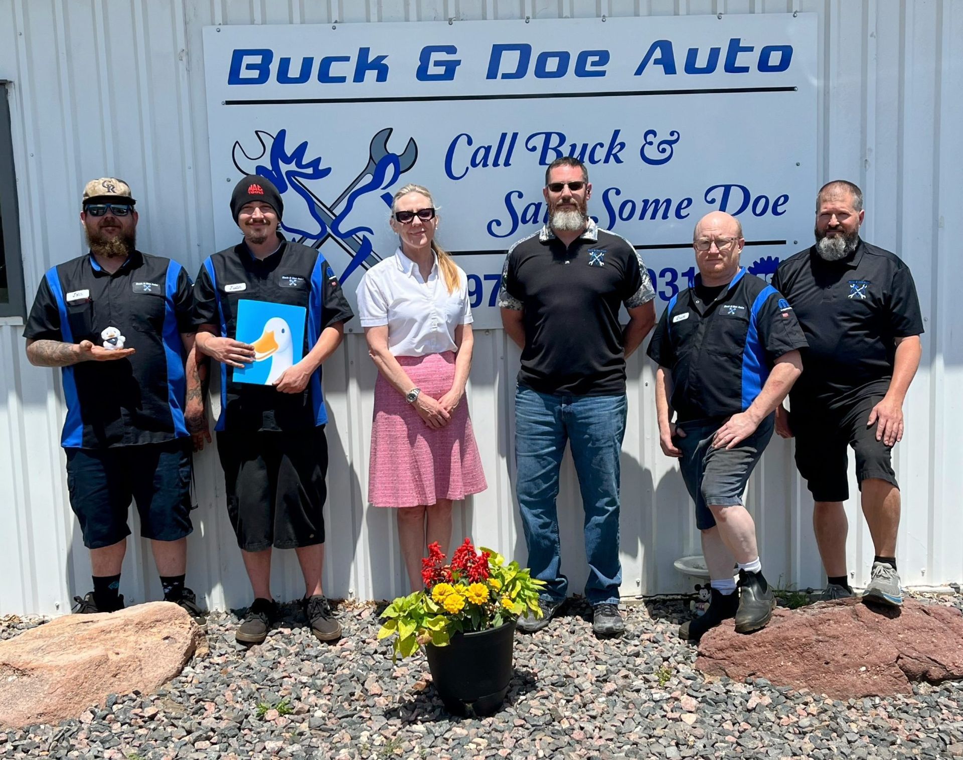 Six people posing outside Buck’s Doe Auto sign in front of a white wall and flower planter. | Buck & Doe Auto