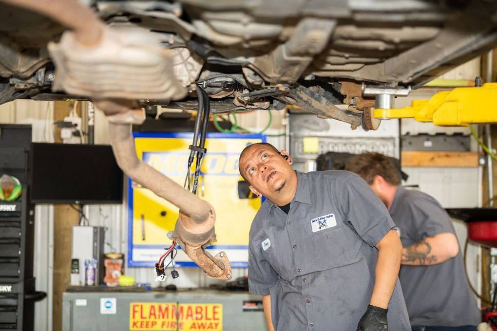 Mechanic inspecting car undercarriage in an auto shop, with a lifted vehicle and machinery overhead. | Buck & Doe Auto