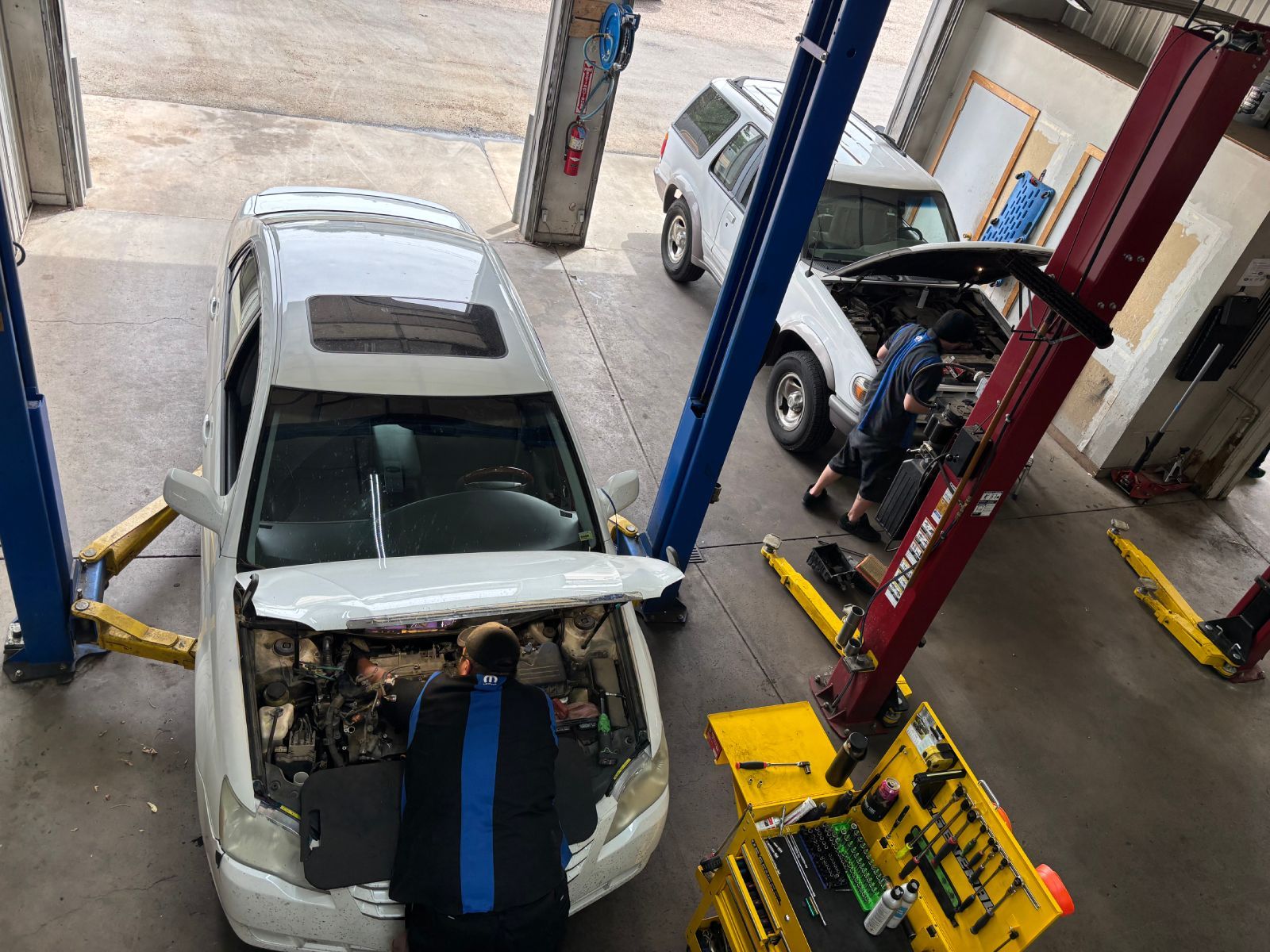 White car in a repair garage, hood open and front bumper removed, with two mechanics nearby. | Buck & Doe Auto