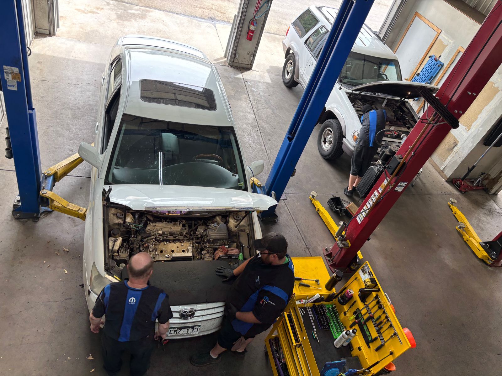 Car in auto repair shop lifted on hoist, mechanics working on front end and engine bay | Buck & Doe Auto