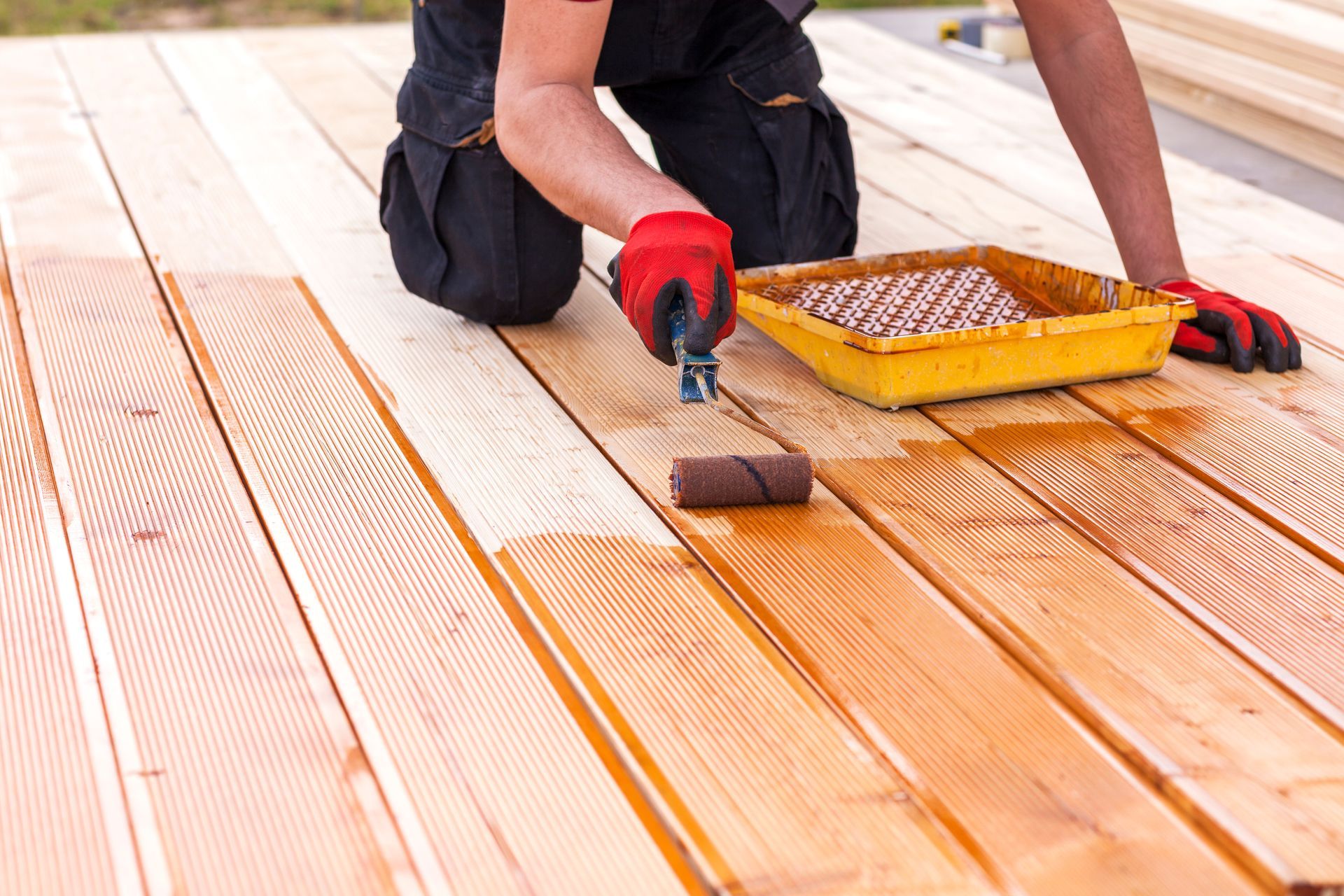 A person wearing gloves kneeling on a wooden deck, applying stain with a paint roller from a yellow tray.