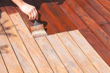 A hand uses a brush to apply a dark brown wood stain to weathered, light-colored wooden deck planks.