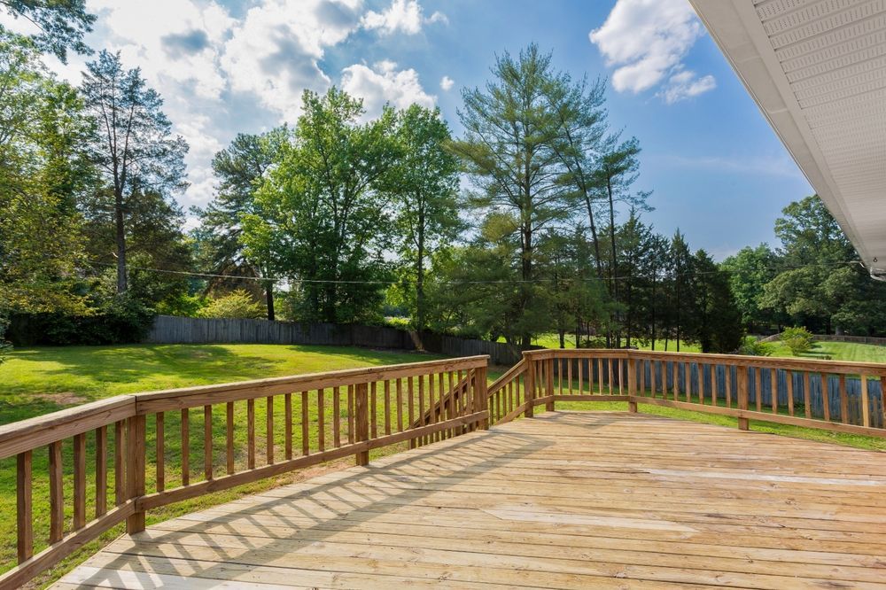 A wooden deck with railings overlooking a large, grassy backyard surrounded by trees under a sunny, blue sky.