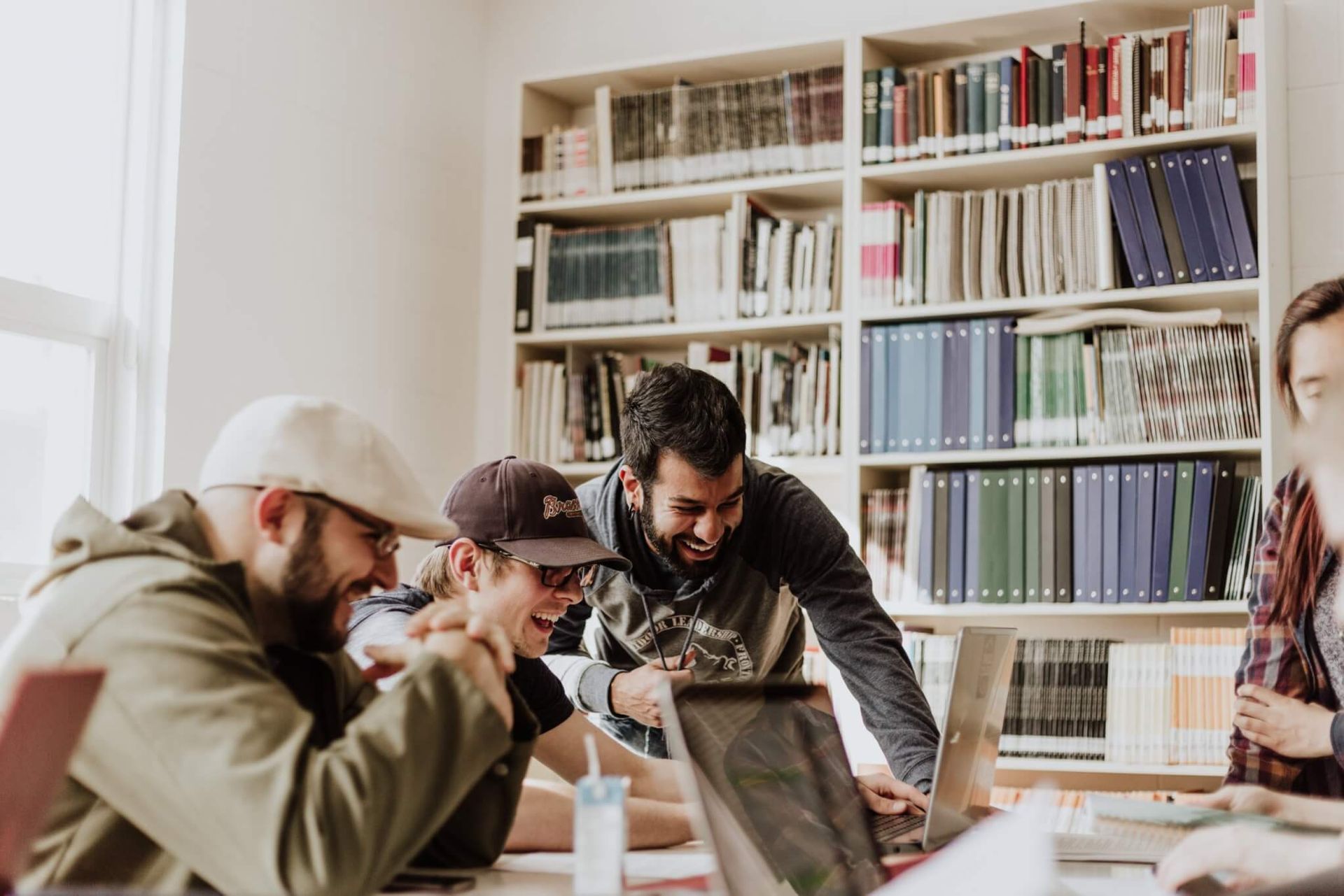 Pessoas colaboram em torno de laptops em uma biblioteca com estantes de livros ao fundo.