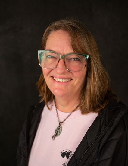 Smiling person with glasses and brown hair in a black jacket against a dark background