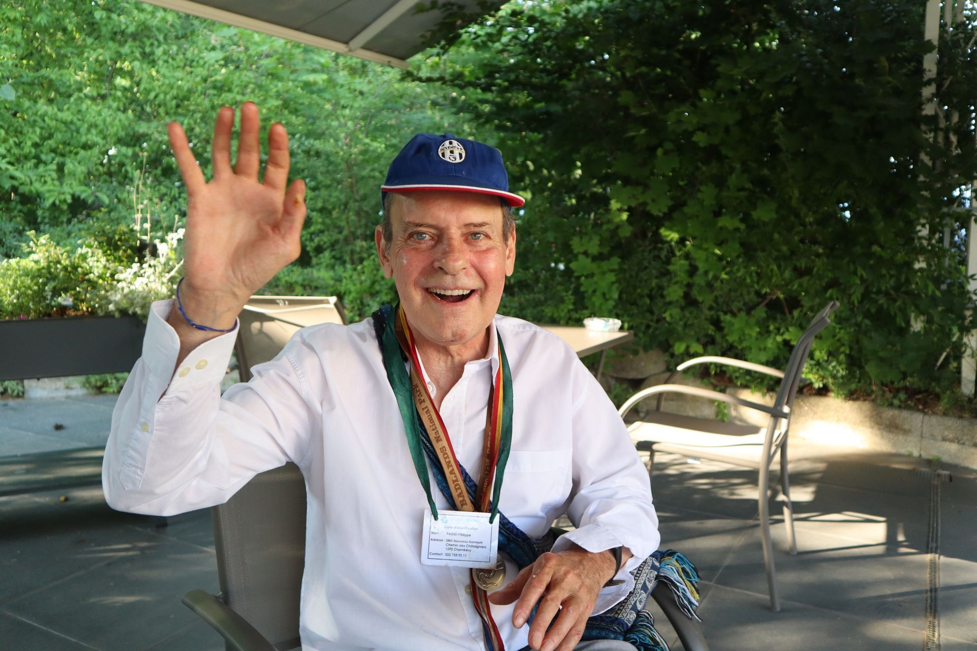 Un homme avec un chapeau bleu est assis sur une chaise et fait signe.