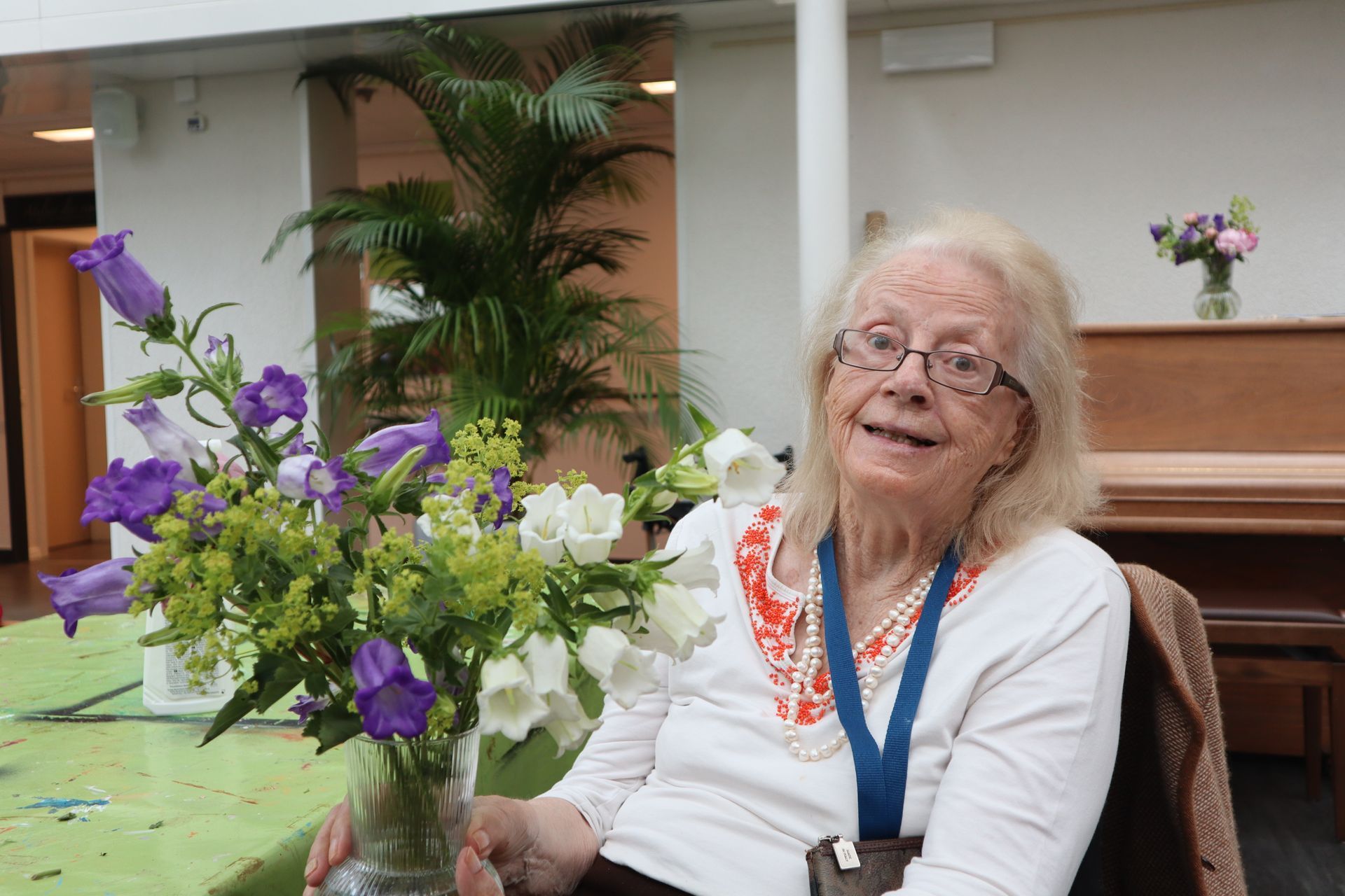 Une femme âgée est assise à une table tenant un vase de fleurs.