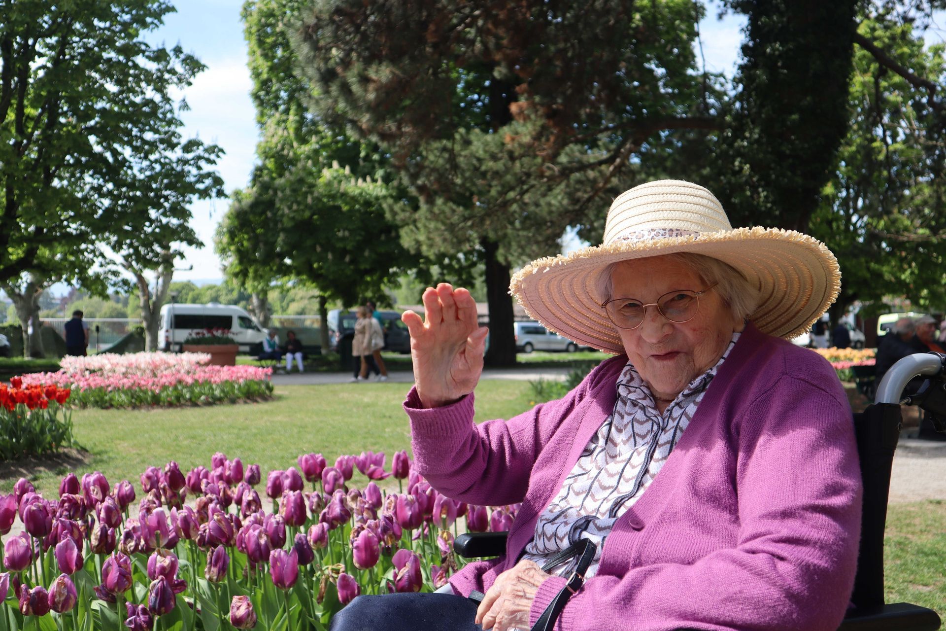 An elderly woman wearing a hat is sitting on a bench in a flower-filled park.