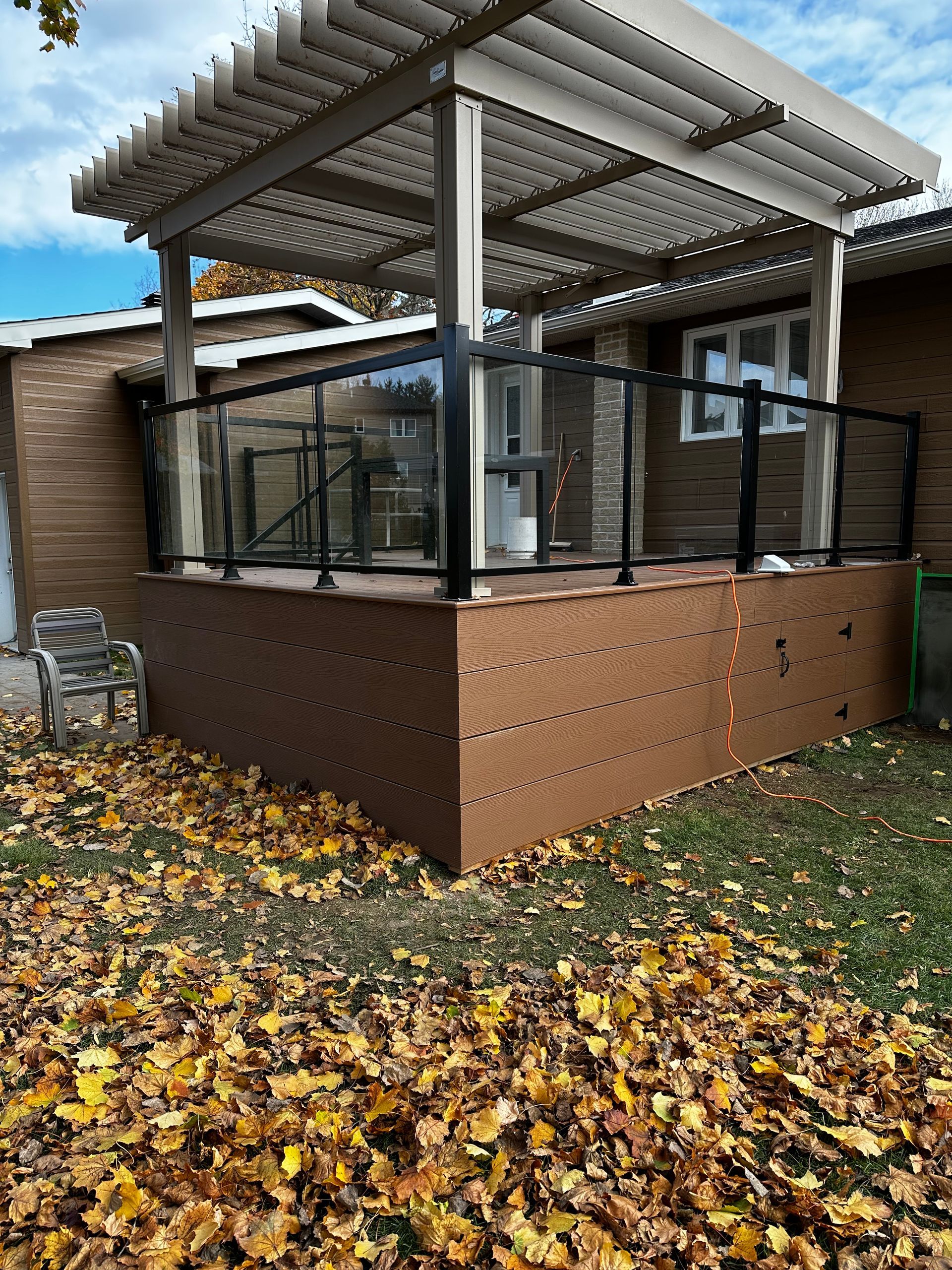 Une terrasse avec une pergola et des feuilles au sol devant une maison.