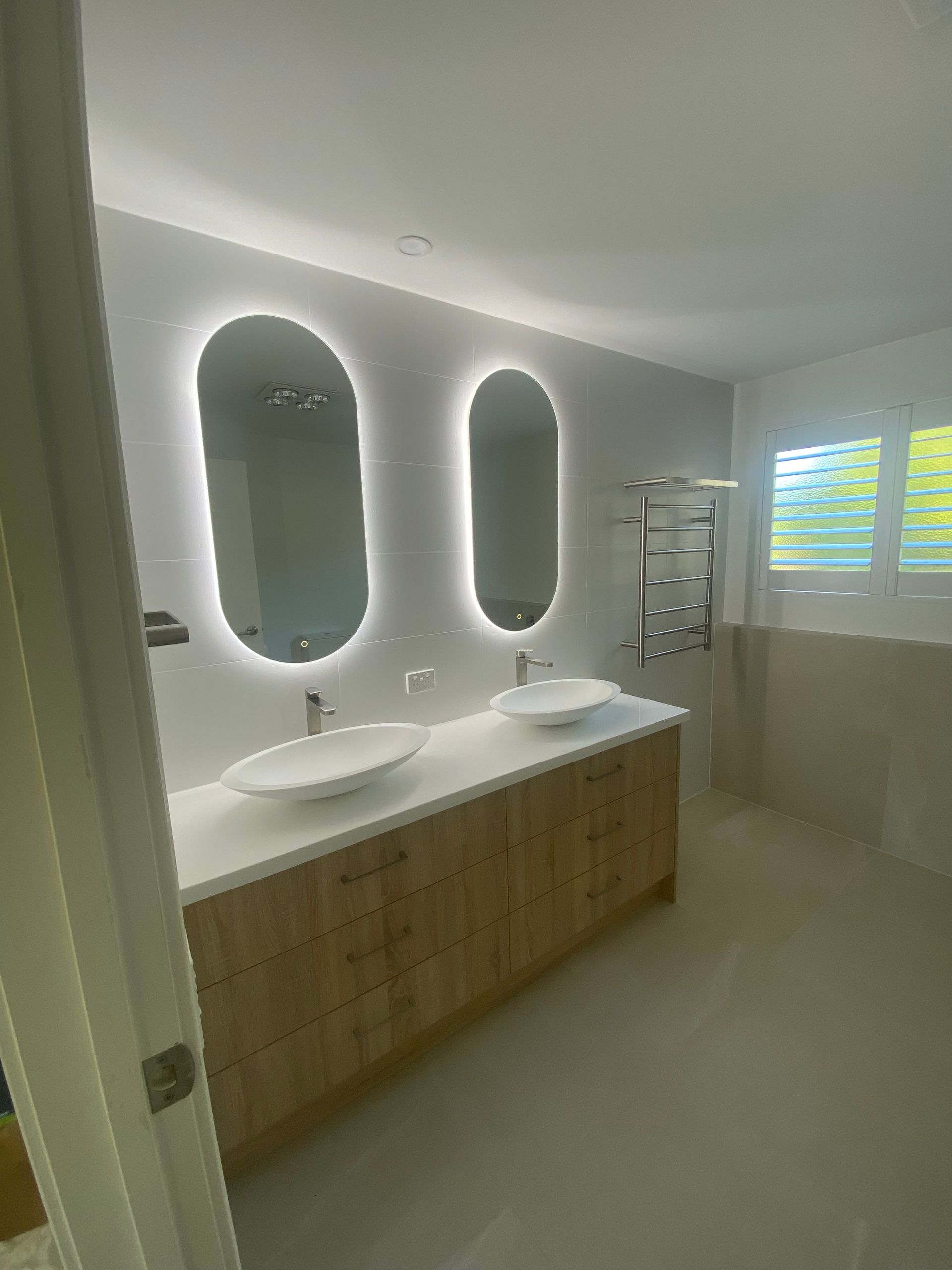 Bathroom with wood vanity, oval backlit mirrors, white sinks, and a towel warmer. — Cut N Edge Panel Factory In Coolum Beach, QLD