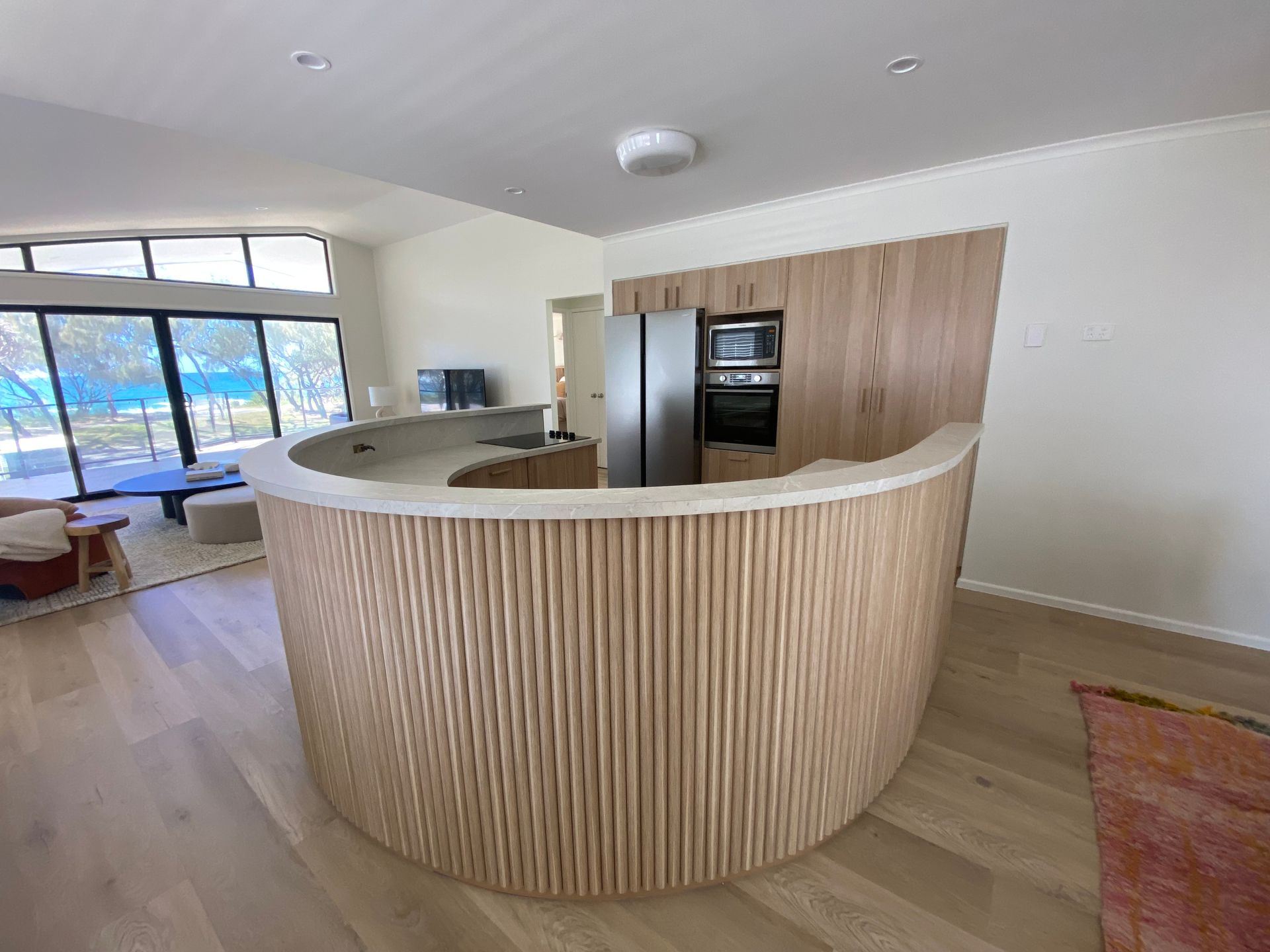 Curved wooden kitchen island with light wood floors and cabinetry, centered in a bright room. — Cut N Edge Panel Factory In Coolum Beach, QLD