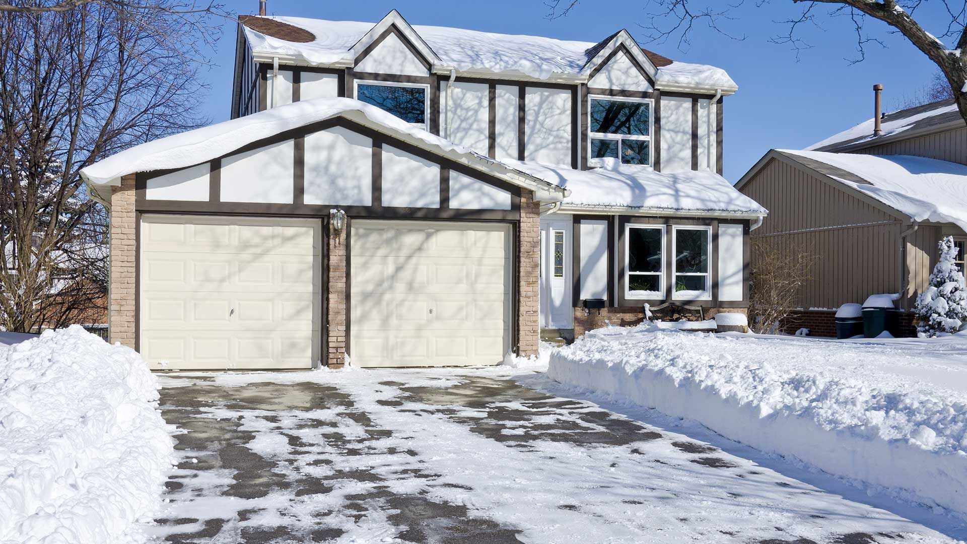 Two-story house with snow-covered roof and driveway; a garage with two doors and a snowy front yard.