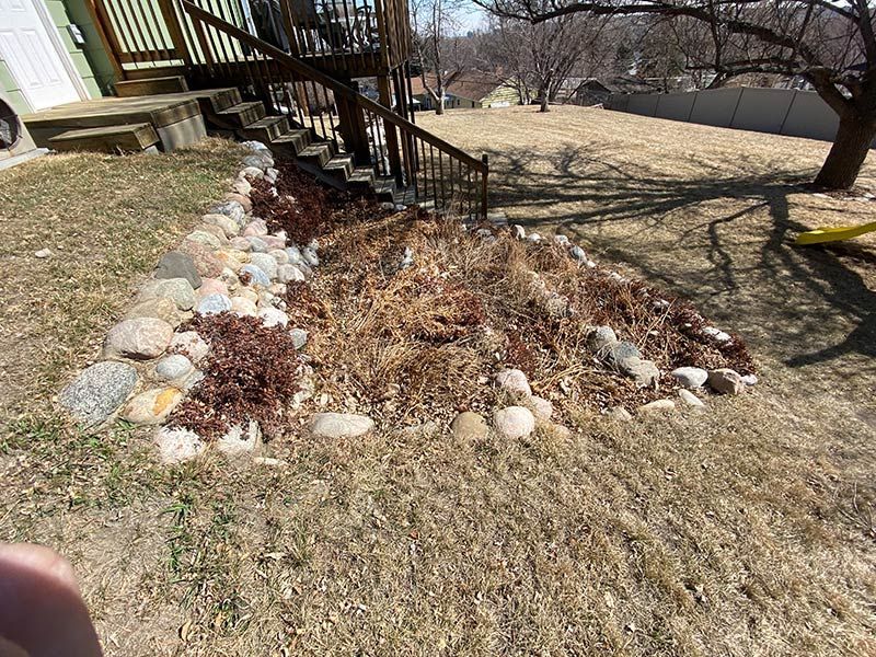 A flower bed with rocks and dead plants is beneath wooden stairs. Dry grass surrounds it.