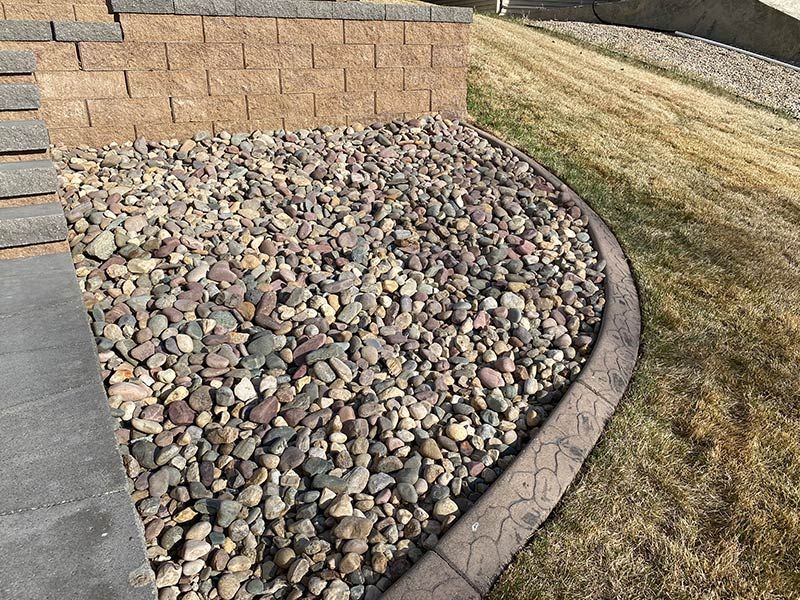Rock bed next to retaining wall and concrete border. Tan and brown rocks and dry grass.