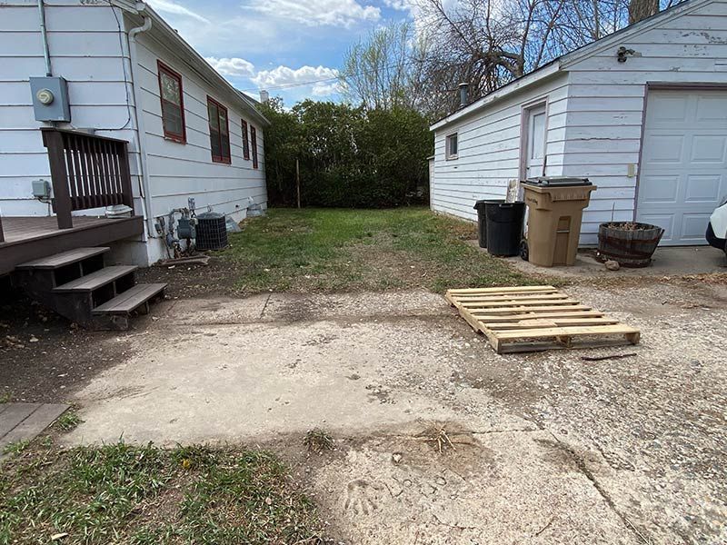 Backyard with houses, garage, wooden steps, pallets, trash cans, and some grass.