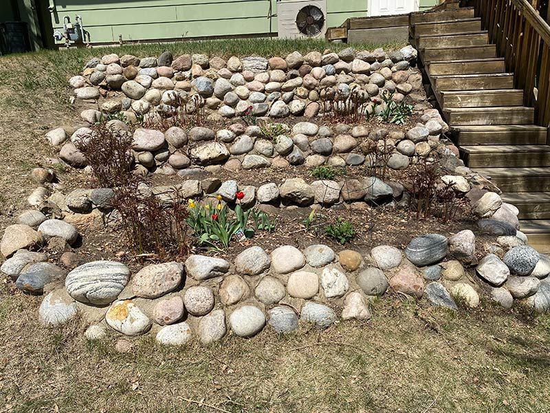 Stone-edged tiered garden bed with emerging spring plants next to wooden stairs.