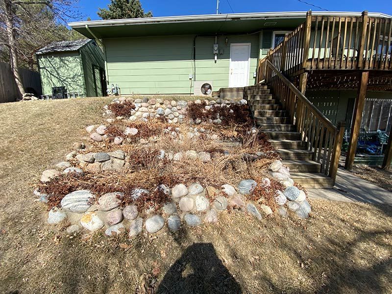 Backyard view: House with green siding, wooden deck, and stone retaining wall. Overgrown, dried plants in the wall.