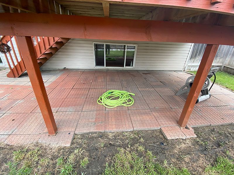 Under a deck, red brick patio with green cord and power washer.