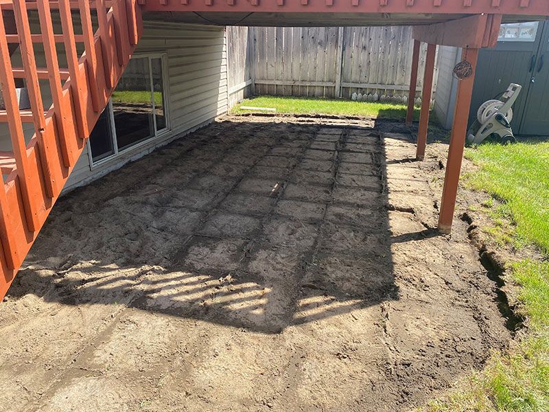 A dirt patio under a deck, ready for paving stones. Grass borders the edge.