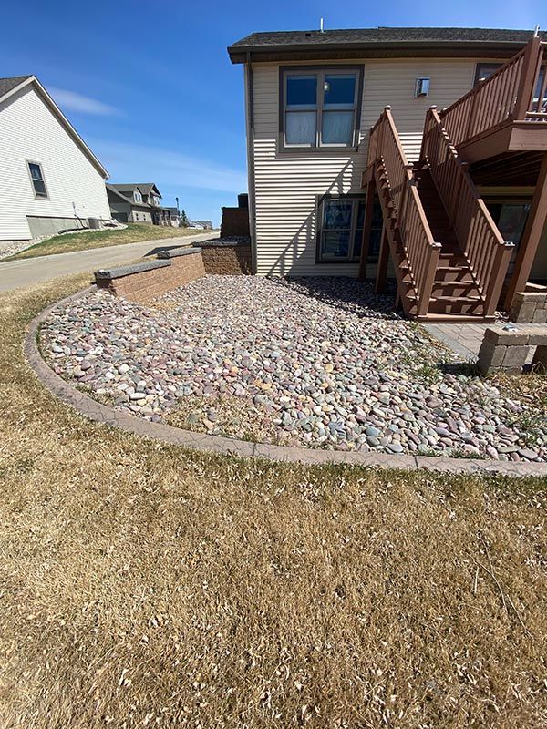 A flower bed with rocks and dead plants under a deck, next to a grassy lawn.