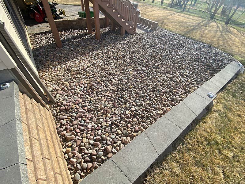Gravel bed beside a house with stairs. Gray retaining wall on the right. Brown and tan rocks.